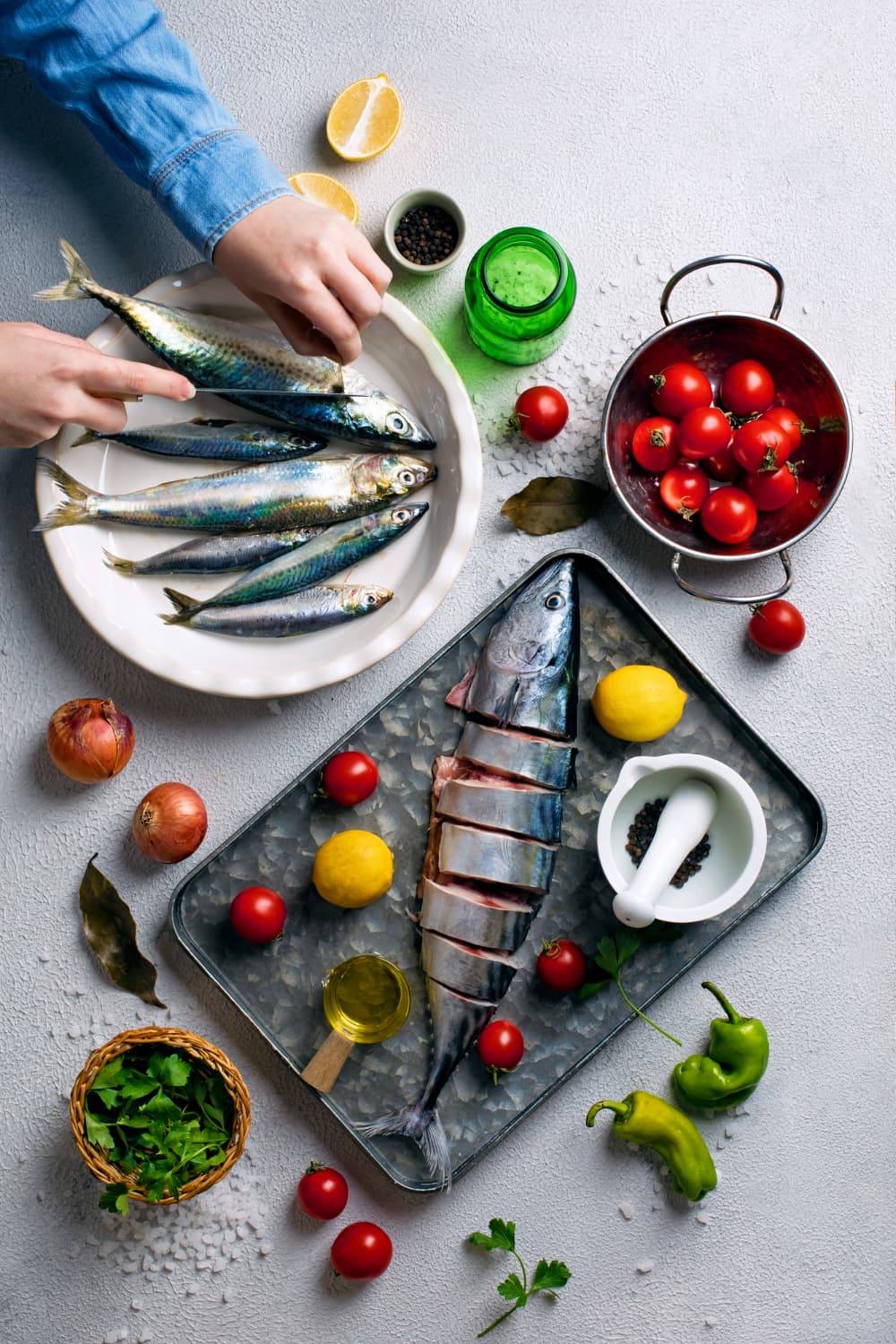 A cook's hands lifting fish from a plate, with a tray of whole fish, tomatoes, herbs and lemon, seen from above