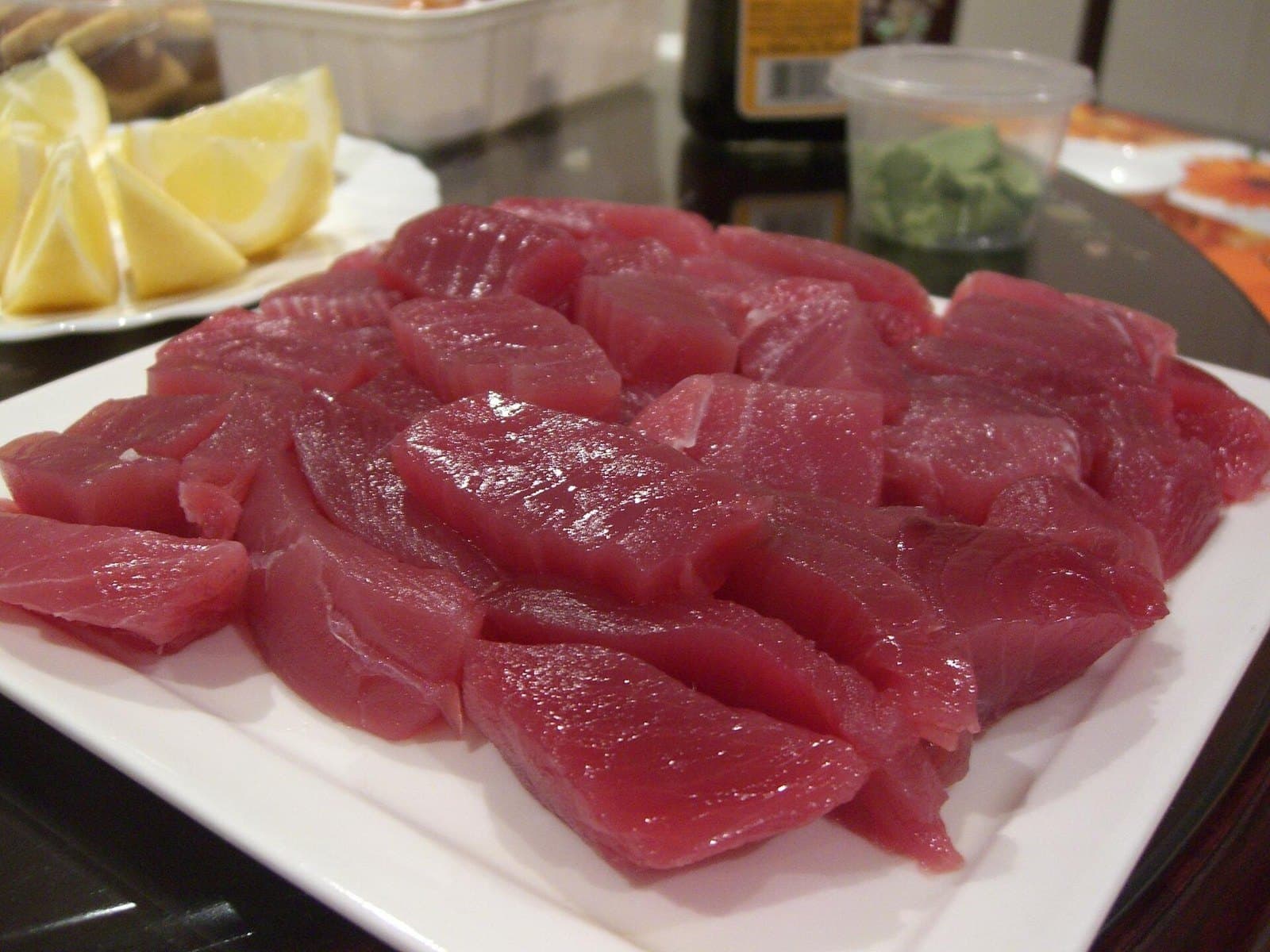 Pieces of raw red tuna cut for sashimi on a white plate, with lemon and wasabi behind