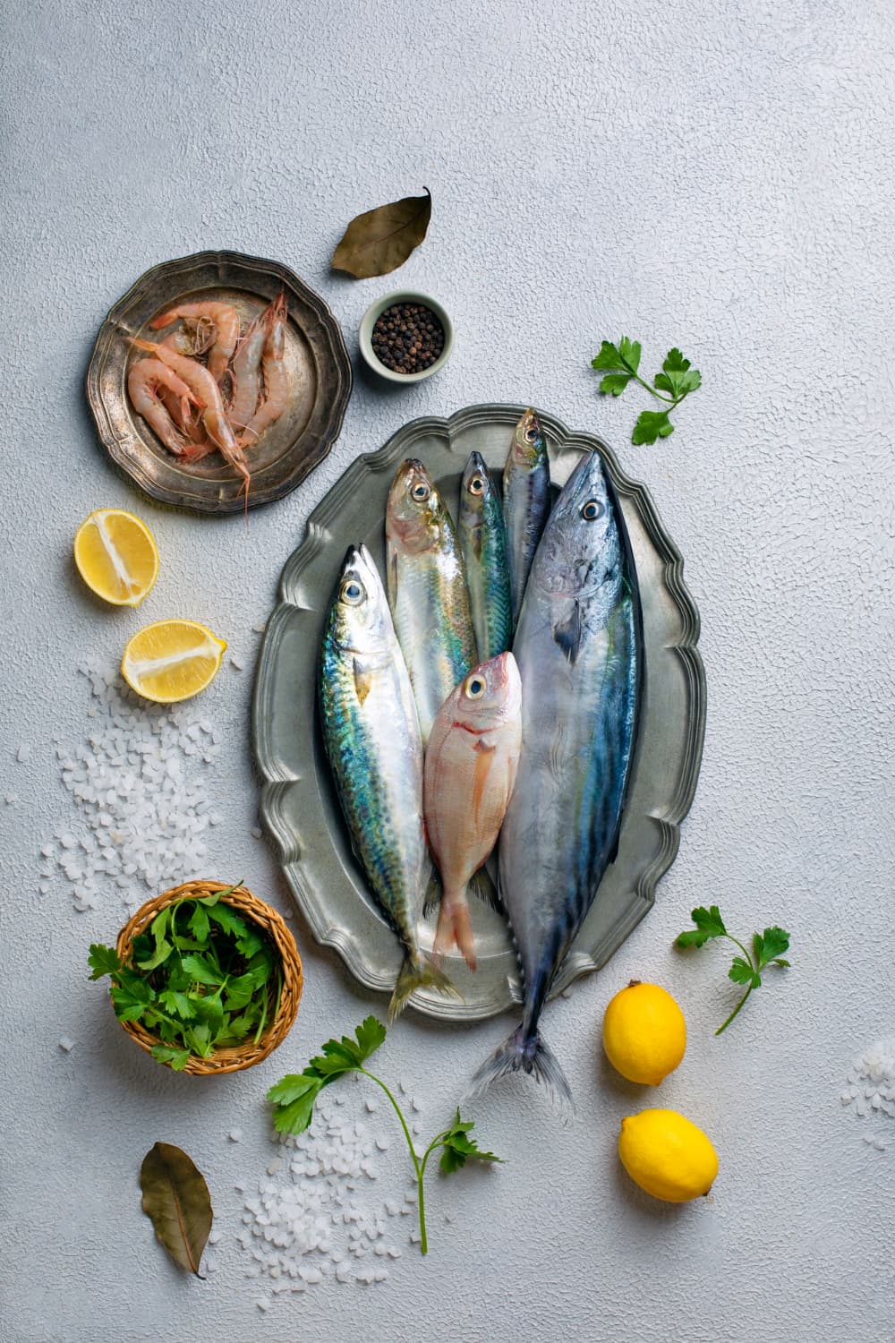 Whole fish on an oval plate with lemon, herbs, salt and a bowl of prawns, on a grey bench